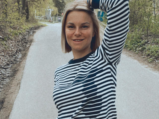 BelleLisaG avec des cheveux blonds, un t-shirt rayé noir et blanc, le bras levé, souriante, debout sur un chemin de gravier, montrant ses seins, dans une zone boisée..