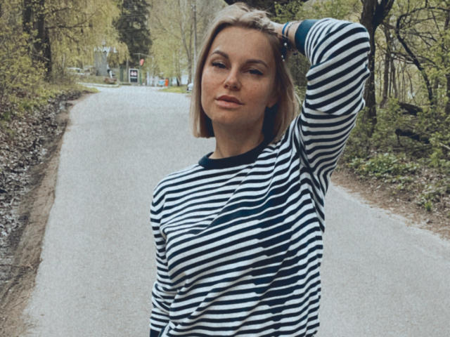 BelleLisaG avec des cheveux blonds, une chemise rayée noire et blanche, une main dans les cheveux, un air confiant, de petites seins, un léger sourire, debout sur une route pavée, avec un arrière-plan de forêt..