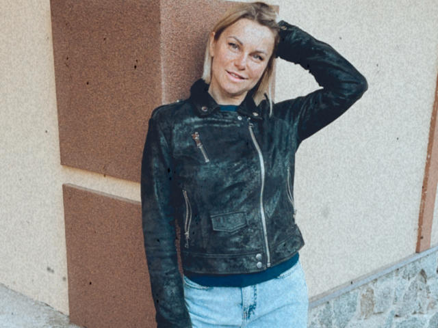 BelleLisaG, avec des cheveux blonds, portant une veste en cuir noir et un jean bleu, debout contre un mur, souriante, une main dans les cheveux, montrant une jolie fesse, look décontracté, plan moyen..