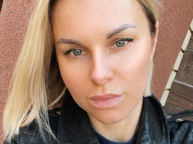 BelleLisaG a des paillettes sur le visage, des yeux verts et des cheveux blonds, elle porte une veste en cuir noir, avec des lèvres légèrement pincées et une expression sérieuse..