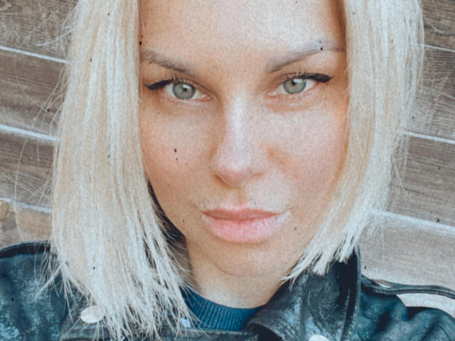 BelleLisaG, avec ses cheveux blonds, ses yeux verts et un léger froncement de sourcils, porte une veste en cuir noir et a quelques petites taches de rousseur sur son visage. Ses lèvres sont légèrement entrouvertes..
