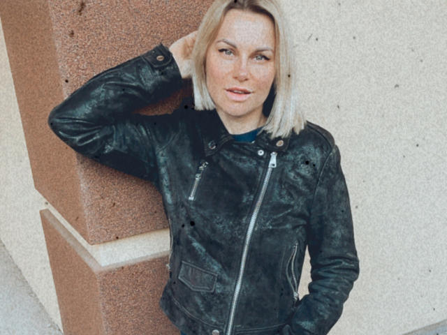 BelleLisaG, avec ses cheveux blonds, ses yeux verts et ses taches de rousseur, portant une veste en cuir noir, debout devant un mur texturé, une main dans la poche, regardant légèrement sur le côté..