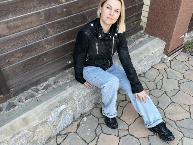 BelleLisaG, avec ses cheveux blonds, sa veste en cuir noir, son jean bleu et ses bottes noires, est assise sur des marches de pierre, les mains posées sur ses genoux, et regarde l'appareil photo. Elle porte des chaussures en cuir noir..
