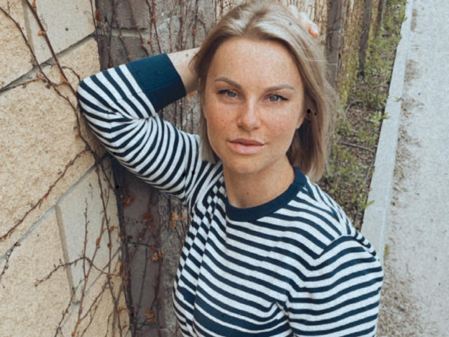 BelleLisaG, avec ses cheveux blonds, portant une chemise rayée noir et blanc, est appuyée contre un mur de briques recouvert de lianes, une main dans ses cheveux, et regarde directement l'appareil photo avec une expression neutre..