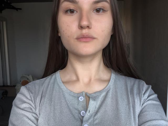 Cathryn Jacquet hat ein ernstes Gesicht, langes braunes Haar, helle Haut und kleine Pickel. Sie trägt ein graues, geripptes Hemd mit Knopfleiste, wobei ihre Brüste nicht sichtbar sind..