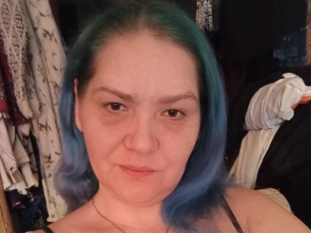 MinnyJeannie tem cabelo azul-esverdeado, pele clara e uma expressão neutra. Ela está usando uma alça preta, e seu nariz é ligeiramente arrebitado. Seus seios são pequenos, e ela está usando um colar..