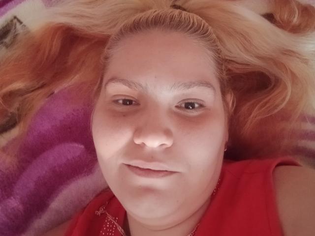 El rostro de MinnyJeannie, con cabello rubio, ojos marrones y una ligera sonrisa, vestida con una camisa roja, acostada sobre una manta morada, mostrando su nariz y una pequeña marca de belleza discreta..