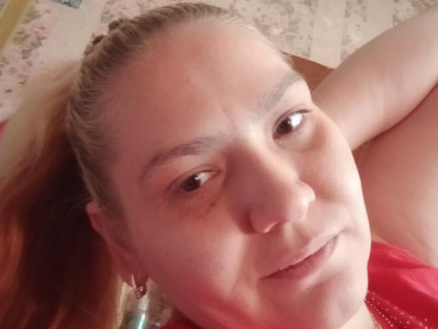 MinnyJeannie, con una coleta rubia, piel clara y una ligera sonrisa, mostrando su rostro y una camisa roja, con sus pequeños senos visibles en la parte superior derecha. Sus ojos marrones miran hacia adelante..