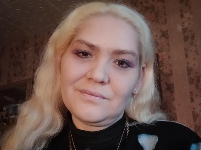 MinnyJeannie a la peau pâle, de longs cheveux blonds, et porte un eye-liner violet avec un rouge à lèvres rose discret. Elle sourit légèrement, et elle a un anneau de nez ainsi que de petites boucles d'oreilles..