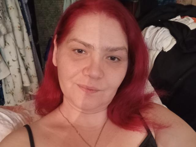 MinnyJeannie, con cabello rojo, piel clara y una ligera sonrisa, vestida con un top negro de tirantes finos, mostrando sus pequeños pezones desnudos y un collar de oro..