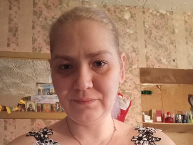 MinnyJeannie heeft blond haar, een lichte huidskleur en kleine borsten. Ze draagt een zwart-wit bloemenblouse en ziet er iets vermoeid uit, met een subtiele glimlach..