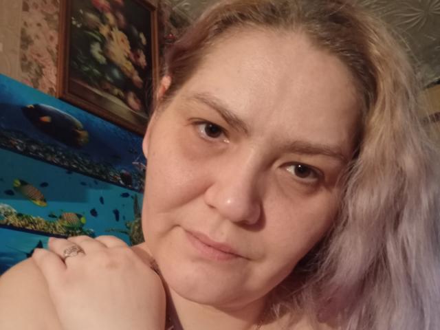 MinnyJeannie mit helllila, leicht zerzausten Haaren, die in die Kamera lächelt, ein kleines, neutrales Lächeln, die ihre Schulter hält, einen Ring an ihrer linken Hand zeigt und braune Augen hat..