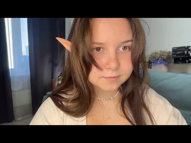 Kathrin Maddoy, com orelhas de elfo, olhos castanhos e cabelos longos castanhos, vestindo uma blusa branca e um colar de prata, tem um rosto adorável e um leve semblante de descontentamento, em um selfie..
