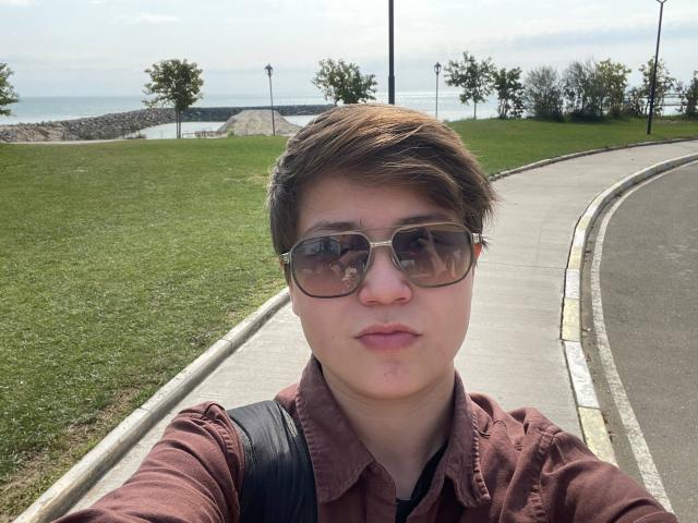 MarybelleDoerr has short brown hair, wears dark sunglasses, a brown shirt, and a black backpack, with a slightly pouty face in a selfie, standing on a curved sidewalk.