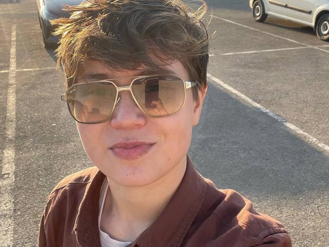 MarybelleDoerr with short brown hair, wearing gold aviator sunglasses and a brown jacket, has pinkish lips and a slight smile, standing in a sunlit parking lot.