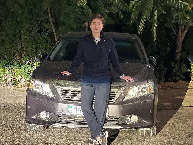MarybelleDoerr, sitting on a dirty brown car with her ass on the hood, wearing a navy hoodie, blue jeans, and white sneakers, has short brown hair and a smile.