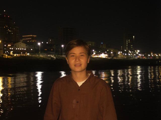 MarybelleDoerr, with short brown hair, smiling at night, wearing a brown button-up shirt, standing by a reflective water surface, city lights in the background.