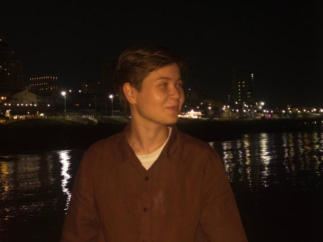 MarybelleDoerr with short brown hair, wearing a brown button-up shirt and white tee, gazing to the side at night, reflection in water, city lights in background.