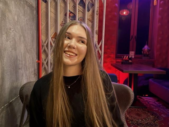 Rochell Dass, met lang bruin haar, lachend, gekleed in een zwarte top, zittend in een stoel, met rode en paarse verlichting, ziet er gelukkig, schattig en aantrekkelijk uit, in een gezellige kamer..