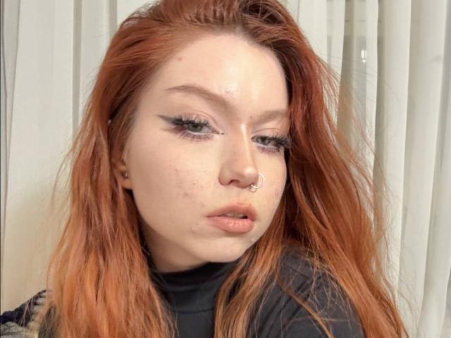 LilliamChevalier has red hair, a nose ring, and a few zits on her face. She's wearing black eyeliner and a black turtleneck. Her lips are slightly parted.