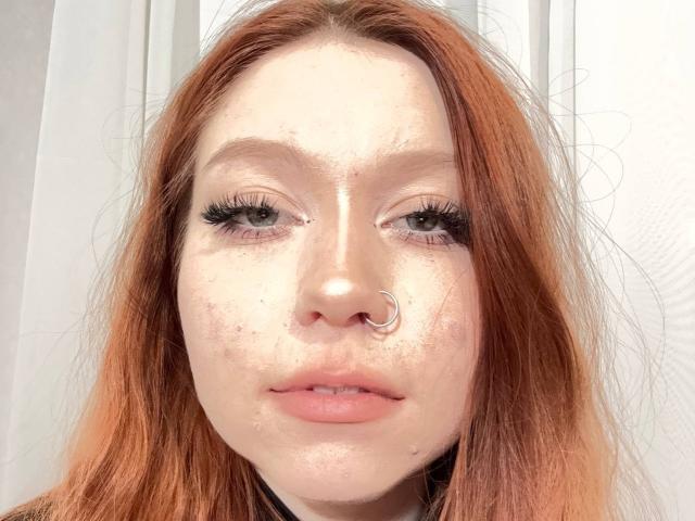 LilliamChevalier has red hair, a silver nose ring, and freckles, with long eyelashes and pink lips, looking directly at the camera with a neutral expression.