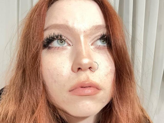 LilliamChevalier has red hair, green eyes with long black eyelashes, a nose ring, and pale freckled skin. Her lips are slightly parted. Her expression is serious and focused.