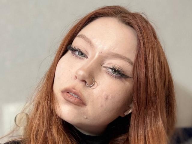 LilliamChevalier has red hair, a nose ring, and light freckles. Her green eyes are half-open with long lashes. She has a slightly pouty face and a few small pimples.