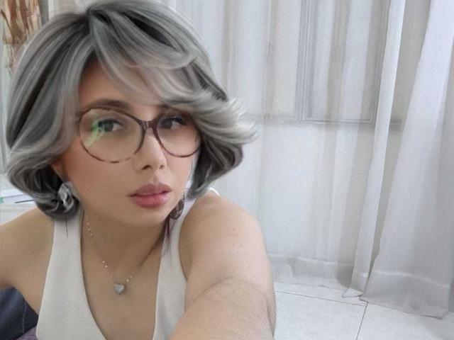 Stephanie Sun, con i capelli grigi e ondulati, occhiali, rossetto rosa, top bianco senza maniche, collana e pelle chiara, guarda la fotocamera, mostrando la parte superiore del corpo e il viso..