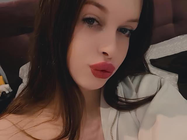 Priscila Nagelhout, com pele clara, cabelo castanho escuro, batom vermelho e delineador preto, vestindo uma jaqueta branca com zíper, olhando para a câmera com um leve semblante de descontentamento..