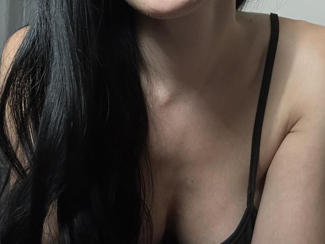 BellaJulls' chest with small to medium tits, black spaghetti straps, long black hair, light skin, and visible collarbone in a low-light, intimate photo.