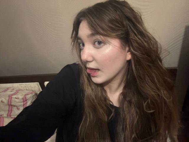 MichikoVivona has long, wavy brown hair, light skin, and is sticking out her tongue while looking at the camera. She's wearing a black top.