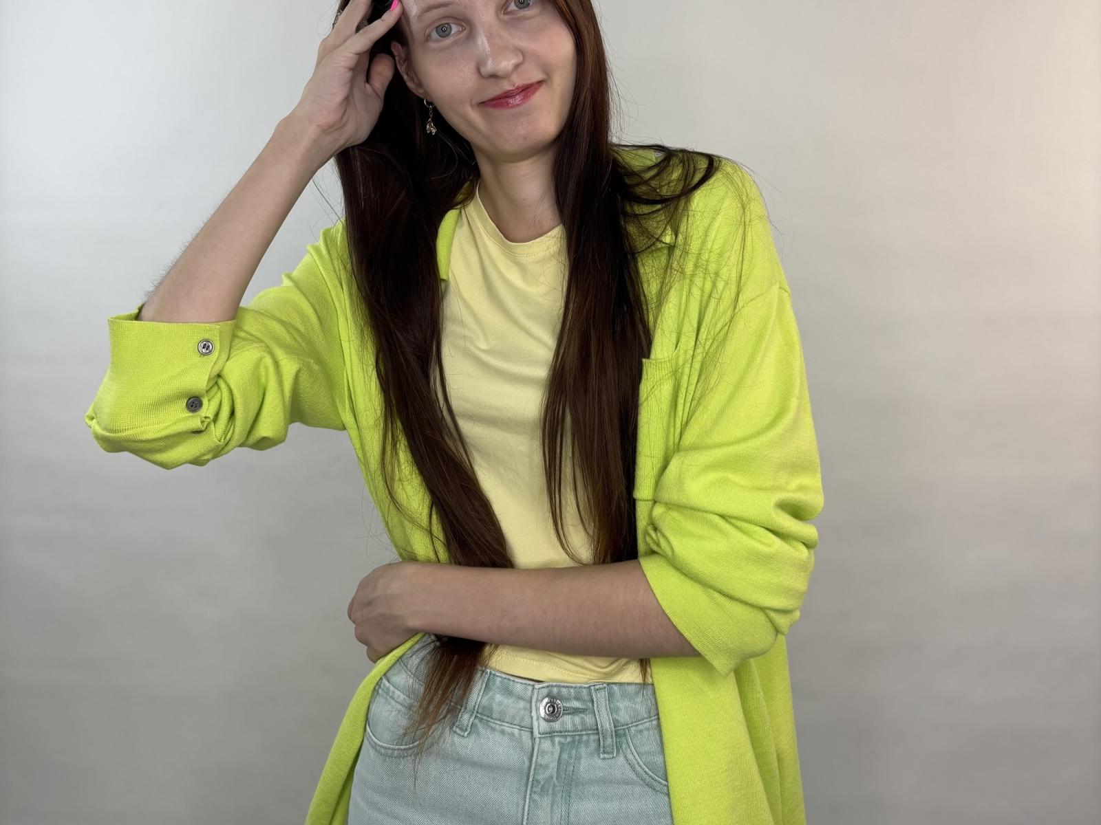 DoriaHoag, con lunghi capelli castani, una camicia gialla e jeans azzurri, sorridente, con una mano tra i capelli, indossa una giacca verde neon, è in piedi e trasmette un'aria carina e rilassata..