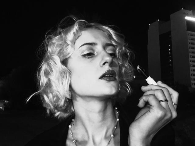Black-and-white photo of MagdalineChrystal with curly blonde hair, applying dark lipstick with a brush, wearing a necklace and ring, looking slightly up with a serious expression.