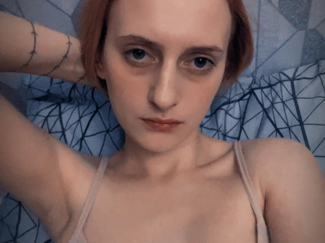 MagdalineChrystal met rood haar, bleke huid, kleine borsten en een tatoeage op haar arm, draagt een beige bh en ligt op een bed met een geometrisch patroon, kijkend in de camera..