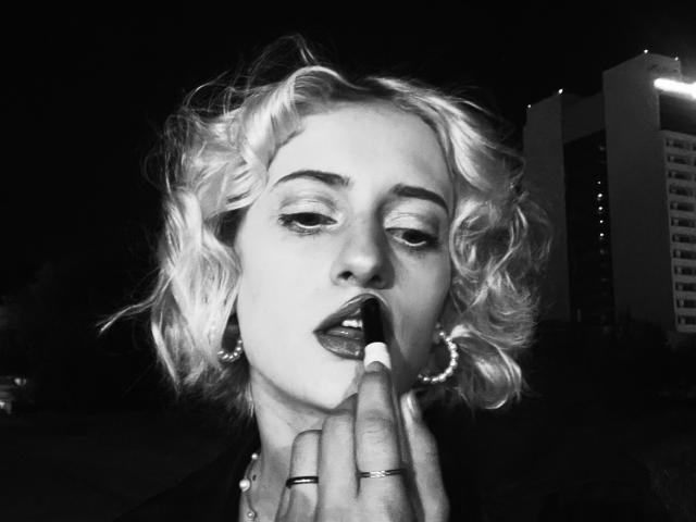 Schwarz-weißes Foto von Magdaline Chrystal mit lockigem, blondem Haar, leicht geöffnetem Mund, Finger im Mund, die Ohrringe und Ringe trägt, Nacht, Stadtlichter im Hintergrund..