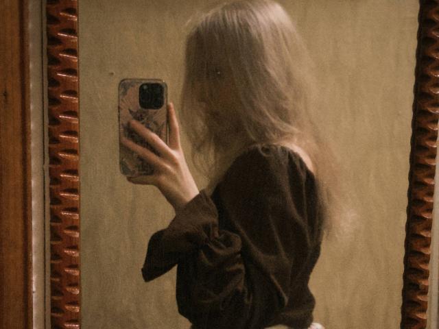 TiffaniMacandog with long, blonde hair, blurry in a brown shirt, taking a selfie in a mirror with a phone, her ass slightly visible. Mirror has a wooden frame.
