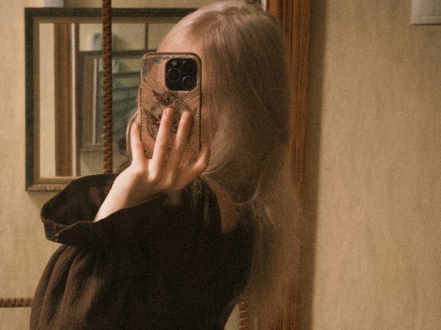 TiffaniMacandog with long blonde hair, wearing a black dress, taking a mirror selfie with her phone, covering her face, showing her hand with a ring.