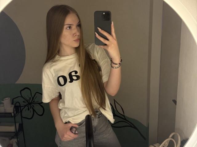 RochellDass with long brown hair, white t-shirt, "06" on chest, gray jeans, holding phone, mirror selfie, slight pout, bracelet, in dimly lit bathroom.
