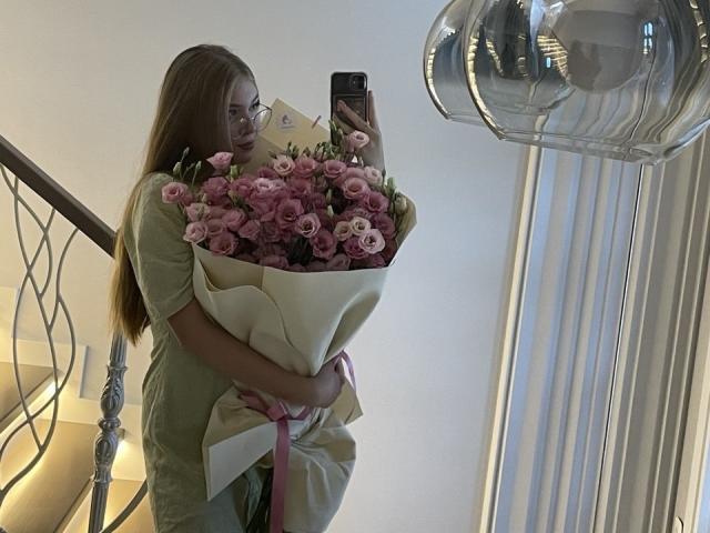 RochellDass, avec de longs cheveux bruns, vêtue de vert, tenant des roses roses dans du papier blanc, prenant un selfie avec son téléphone noir, debout près d'une rambarde en métal et d'une lampe argentée..