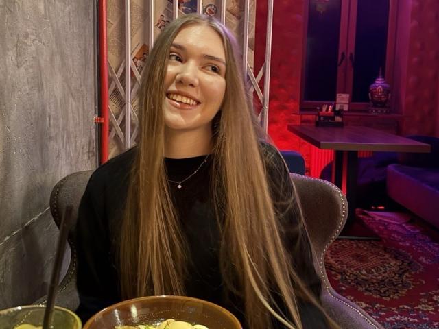 RochellDass with long, straight brown hair, smiling, wearing a black sweater, sitting in a dark room with a red glow, holding a bowl of food in front of her.