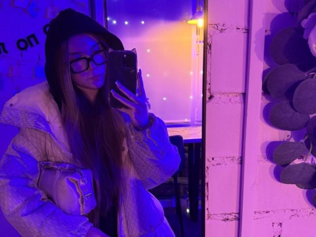 Rochell Dass, met lang bruin haar, zwarte bril en een zwarte hoed, maakt een spiegelzelfportret in paars-blauw licht, gekleed in een pluizige witte jas en met een grijze handtas..