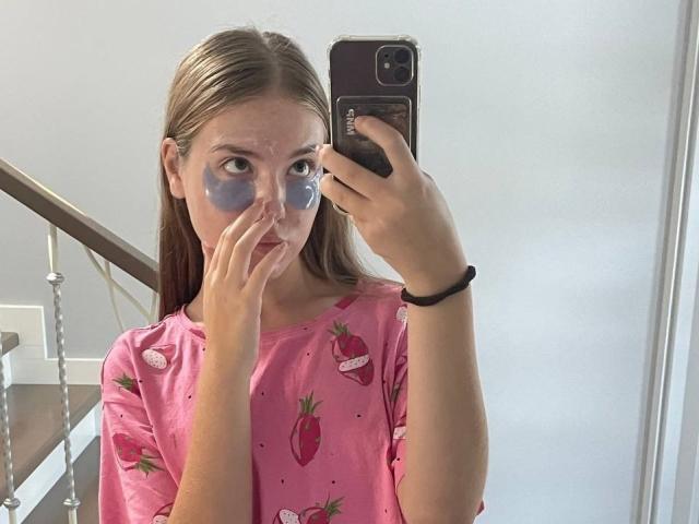 RochellDass with blue face paint, wearing a pink shirt with red strawberry patterns, taking a mirror selfie with an iPhone. Her hand touches her nose.