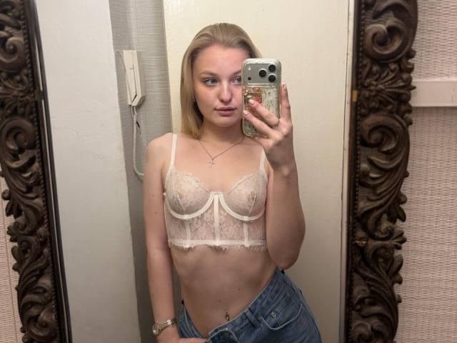 NievesMegee with blonde hair, wearing a white lace bra that shows her tits and a blue denim skirt, taking a mirror selfie with a gold phone.