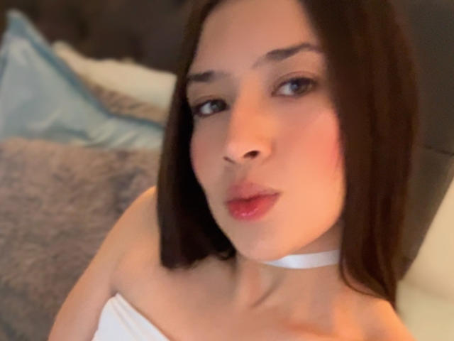 EmmaLombardi with straight brown hair, white strapless top, and white choker, showing pink lips and slightly flushed cheeks, looking at camera with soft, sexy expression.