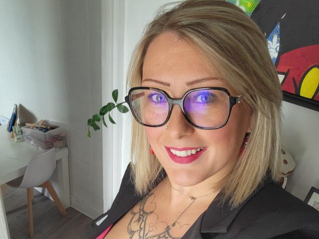 AmandalaFrancaise with blonde hair, black glasses, red lipstick, nose piercing, and necklace, smiling at camera, showing blue light reflection in glasses, wearing black shirt.