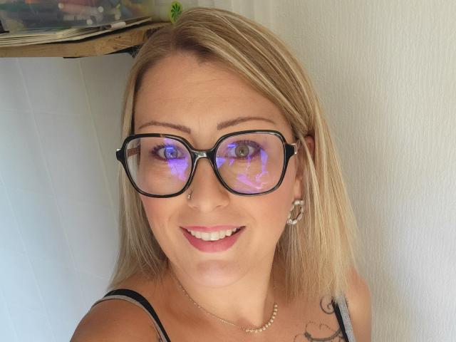AmandalaFrancaise with blonde hair, black glasses reflecting blue light, nose piercing, smiling, black strap top, tattoo on right shoulder, white wall background, silver hoop earrings.