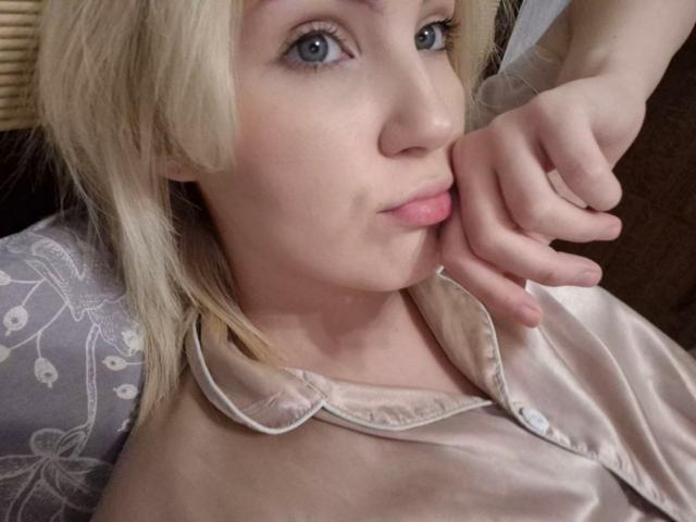 LidiaOckimey has blonde hair, blue eyes, and pink lips. She's wearing a satin shirt, resting her chin on her hand. Her nipples are slightly visible under the shirt.