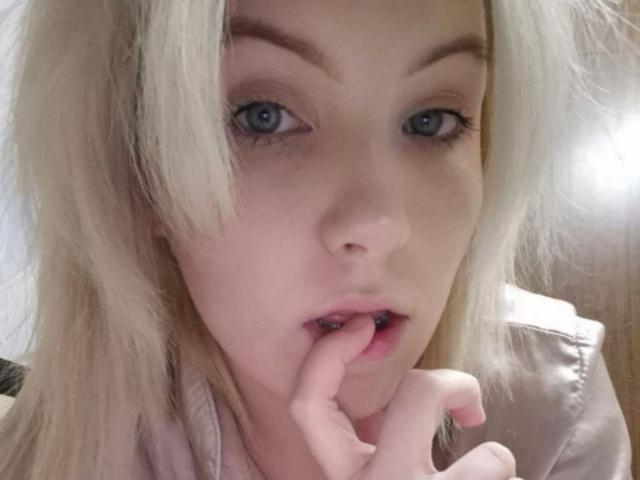 LidiaOckimey with blonde hair, blue eyes, and pink lips, biting her finger with a slightly open mouth, wearing a light pink top, looking seductive.