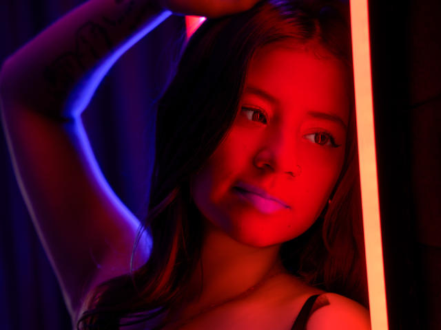 EmmaFeri com iluminação vermelha e azul, braços levantados, mostrando tatuagem no braço direito, usando alça preta, batom roxo, olhando para a câmera com um leve sorriso, cabelo castanho ondulado..