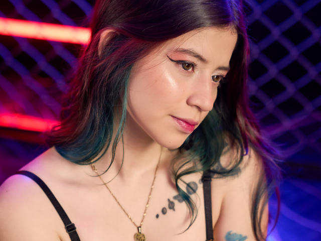 EmmaFeri has teal and black hair, pink winged eyeliner, and a subtle smile, wearing a black strap top with tattoos on her chest, in a colorful, neon-lit setting.